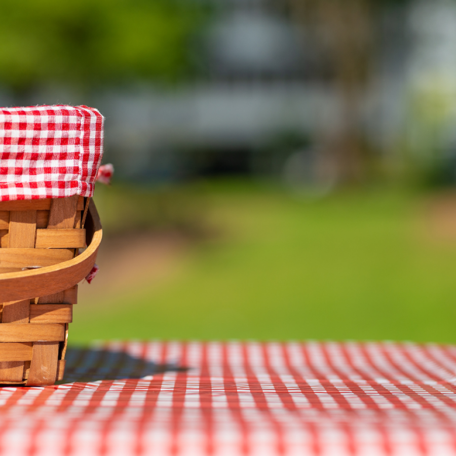 A picnic basket with a red checked cloth over it sitting on a red chequered picnic blanket on a sunny day